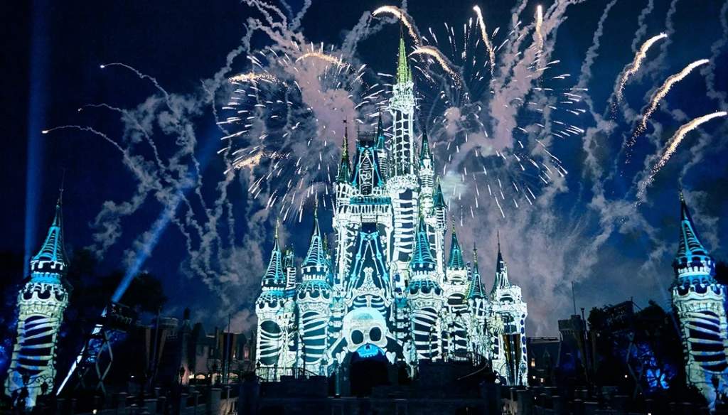 Disney castle lit up with a skull motif projection as fireworks explode overhead at night.